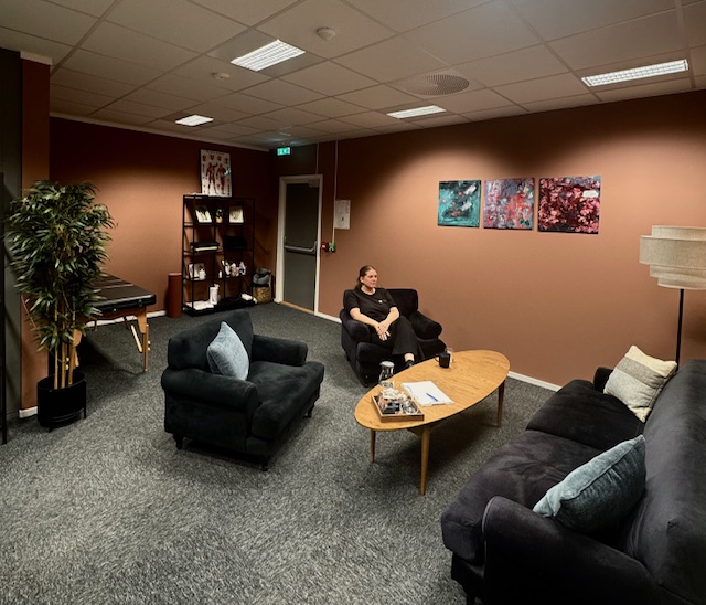 Terapirom hos SansHera Helhetsterapi på Jessheim, to sorte stoler og en sort sofa, salongbord med vann og glass, en terapeut sitter i ene stolen