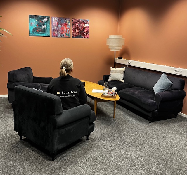 Terapirom hos SansHera Helhetsterapi på Jessheim, to sorte stoler og en sort sofa og et salongbord med vann, en terapeut med SansHera-genser sitter med ryggen til