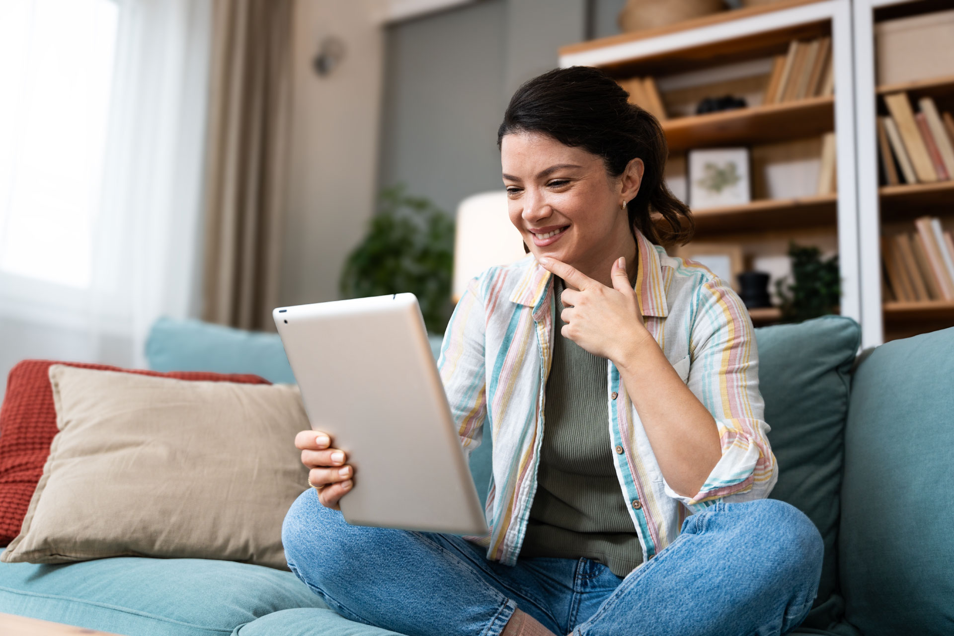 En kvinne sitter i sofaen hjemme og holder et nettbrett. Hun har en online konsultasjon med en terapeut fra SansHera